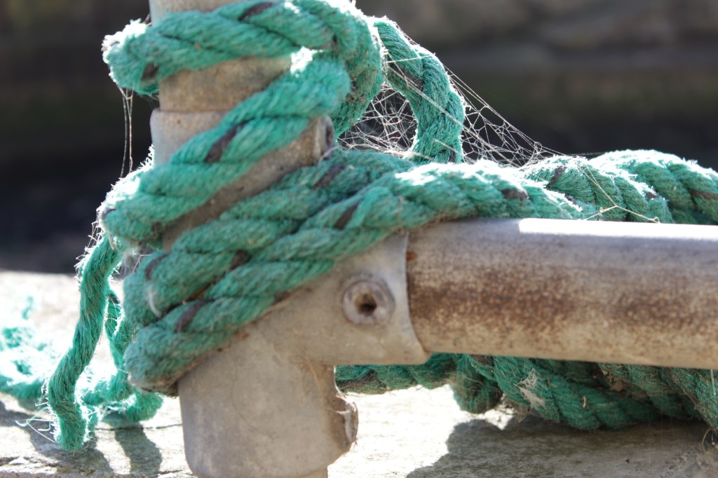 Image of a turquoise fishing rope tied around a metal pole with spider webs catching the sunlight
